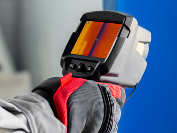 Technician wearing protective gloves holding a handheld thermal imaging camera displaying a heat map.