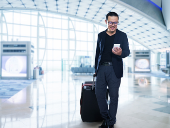 “Man standing in an airport terminal with a rolling suitcase, smiling while looking at his smartphone.