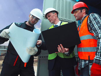 Three workers wearing hard hats and safety gear reviewing blueprints and a laptop at an industrial worksite.