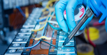 Technician wearing blue gloves soldering electrical connections on a battery pack or electronic circuit.