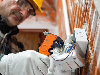 Electrician wearing protective gloves and a hard hat working on exposed wiring inside a wall-mounted electrical box.